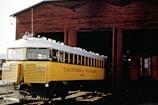 A vintage yellow railcar labeled 'California Western M-100' is partially emerging from an old wooden train shed. The railcar features a white roof and grill, and is positioned on railway tracks in front of the rustic brown structure.