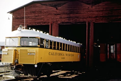 A vintage yellow railcar labeled 'California Western M-100' is partially emerging from an old wooden train shed. The railcar features a white roof and grill, and is positioned on railway tracks in front of the rustic brown structure.