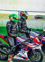 Two motorcyclists wearing colorful helmets and black riding suits are sitting on sport motorcycles. The motorcycles are decorated with red, blue, and white patterns and various logos. They appear to be on a racetrack, given the presence of helmet visors and branded racing gear.