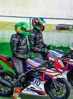 Two motorcyclists wearing colorful helmets and black riding suits are sitting on sport motorcycles. The motorcycles are decorated with red, blue, and white patterns and various logos. They appear to be on a racetrack, given the presence of helmet visors and branded racing gear.