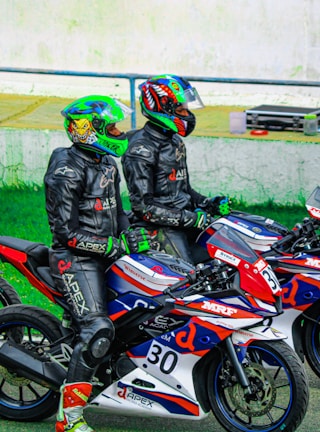 Two motorcyclists wearing colorful helmets and black riding suits are sitting on sport motorcycles. The motorcycles are decorated with red, blue, and white patterns and various logos. They appear to be on a racetrack, given the presence of helmet visors and branded racing gear.