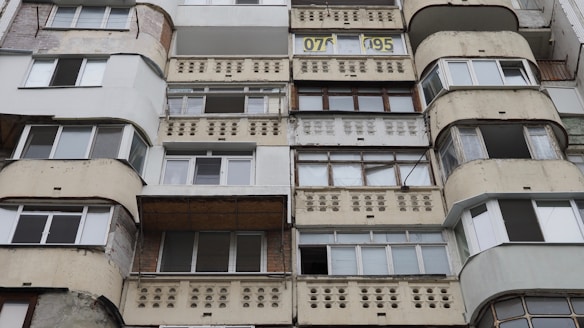 The image features a facade of a multi-story residential building with a repetitive pattern of windows and balconies. The balconies have a distinct design with circular cutouts in the railing. The structure appears worn, with signs of aging on the exterior surfaces. The building has a mix of rectangular and curved elements, with various window types and some sections that look patched or renovated.