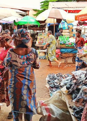 A bustling outdoor market scene with colorful umbrellas providing shade. Several people are shopping and interacting, wearing vibrant, patterned clothing. Various goods, including shoes and packaged products, are displayed on tables and carts.