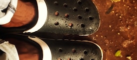 A pair of black slip-on shoes with perforations, possibly Crocs, are seen from above. The shoes are wet, suggesting they have been in contact with water. The ground appears muddy, with scattered small pieces of debris and a hint of green vegetation.