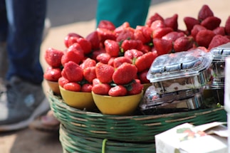 A basket filled with vibrant red strawberries is displayed alongside small plastic containers of the fruit. The strawberries are placed in yellow bowls, and the scene suggests an outdoor market or fruit stall setting. The focus is on the ripe and juicy appearance of the strawberries.