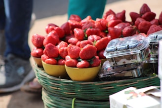 A basket filled with vibrant red strawberries is displayed alongside small plastic containers of the fruit. The strawberries are placed in yellow bowls, and the scene suggests an outdoor market or fruit stall setting. The focus is on the ripe and juicy appearance of the strawberries.