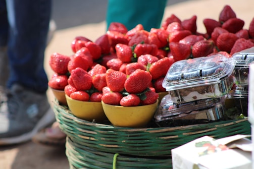 A basket filled with vibrant red strawberries is displayed alongside small plastic containers of the fruit. The strawberries are placed in yellow bowls, and the scene suggests an outdoor market or fruit stall setting. The focus is on the ripe and juicy appearance of the strawberries.