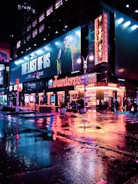 A vibrant city street scene at night, featuring neon lights reflecting on the wet pavement. Large billboards and signs light up the area, including an advertisement for 'The Last of Us'. The street is bustling with people, some sitting at outdoor tables by the restaurant named 'Juniors'.