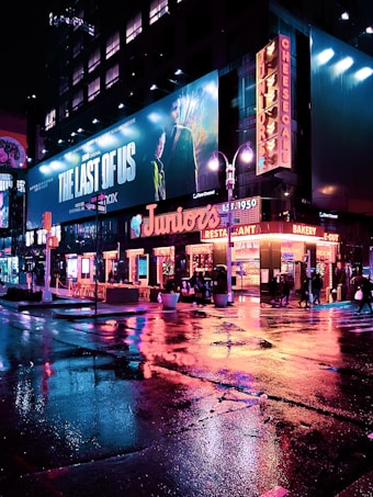A vibrant city street scene at night, featuring neon lights reflecting on the wet pavement. Large billboards and signs light up the area, including an advertisement for 'The Last of Us'. The street is bustling with people, some sitting at outdoor tables by the restaurant named 'Juniors'.