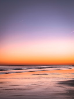 A serene beach landscape with calm waters reflecting the vibrant colors of a sunset. The sky transitions from a soft orange near the horizon to a gentle purple, creating a peaceful and harmonious atmosphere.