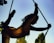 A silhouetted figure with curly hair is performing an acrobatic pose on a rope against a clear sky, with one leg extended gracefully behind. The background shows blurred green foliage.