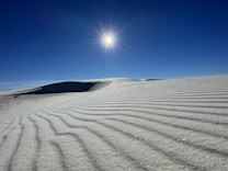 A serene desert landscape with rippled sand dunes stretching into the distance under a clear blue sky. The sun shines brightly overhead, casting sharp shadows across the sand. The scene is devoid of people, emphasizing the tranquil and untouched nature of the environment.