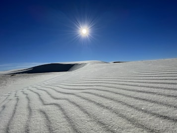 A serene desert landscape with rippled sand dunes stretching into the distance under a clear blue sky. The sun shines brightly overhead, casting sharp shadows across the sand. The scene is devoid of people, emphasizing the tranquil and untouched nature of the environment.