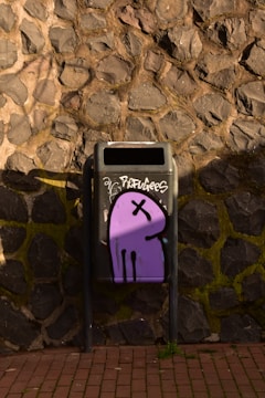 A public trash bin against a stone brick wall is decorated with a unique piece of graffiti. The graffiti features a simple, cartoonish purple figure with a black outline and a cross mark for its eye. The word 'refugees' is written above the artwork in a tag-like style.