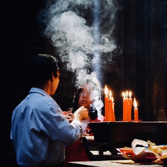 Two people are engaged in a spiritual or religious ritual involving lit red candles. Smoke rises in intricate patterns between them, creating a mystical atmosphere. The setting appears to be dimly lit, focusing on the interaction and the glow of the candles.