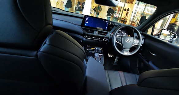 Interior view of a luxury car, featuring a sleek dashboard with a digital display and steering wheel adorned with the Lexus logo. The seats and interior design are modern and sophisticated, showcasing black leather upholstery. Exterior view through the car windows reveals a bustling street scene with pedestrians and storefronts.