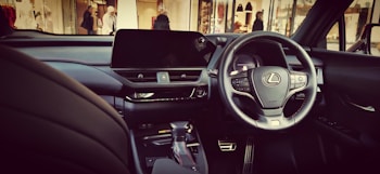 A luxury car interior featuring a sleek black dashboard with a prominent steering wheel displaying the Lexus logo. The car is equipped with modern technology, including a large central display screen and various control buttons. The environment outside the car shows a mall or shopping area with a few people in the background.