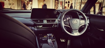 A luxury car interior featuring a sleek black dashboard with a prominent steering wheel displaying the Lexus logo. The car is equipped with modern technology, including a large central display screen and various control buttons. The environment outside the car shows a mall or shopping area with a few people in the background.