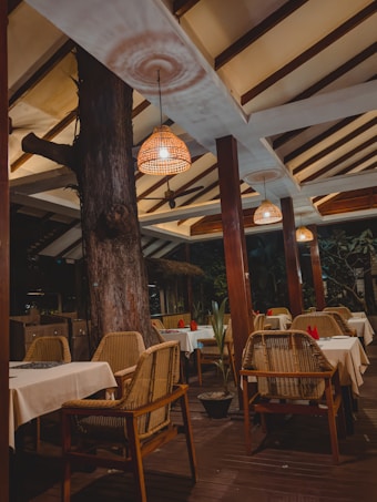 A cozy outdoor dining area with wooden chairs and tables covered in white tablecloths. Woven pendant lights hang from the ceiling, casting a warm and inviting glow. A large tree trunk is integrated into the space, adding a natural element.