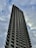 A tall, modern skyscraper extends into a cloudy sky, featuring a distinct pattern of vertical lines and rows of windows. The building has a robust and angular architectural design, creating a striking silhouette against the overcast backdrop.