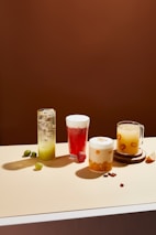 Four different beverages are arranged on a light-colored tabletop against a solid brown background. From left to right, there is a tall glass with a green drink and ice, a glass of red liquid with foam on top and a strawberry beside it, a creamy-looking beverage with orange chunks in a clear glass, and a mug with a yellow drink containing visible slices of fruit. Small fruits and nuts are scattered around the drinks.
