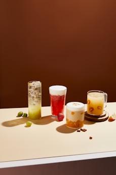 Four different beverages are arranged on a light-colored tabletop against a solid brown background. From left to right, there is a tall glass with a green drink and ice, a glass of red liquid with foam on top and a strawberry beside it, a creamy-looking beverage with orange chunks in a clear glass, and a mug with a yellow drink containing visible slices of fruit. Small fruits and nuts are scattered around the drinks.