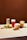 Four different beverages are arranged on a light-colored tabletop against a solid brown background. From left to right, there is a tall glass with a green drink and ice, a glass of red liquid with foam on top and a strawberry beside it, a creamy-looking beverage with orange chunks in a clear glass, and a mug with a yellow drink containing visible slices of fruit. Small fruits and nuts are scattered around the drinks.
