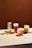 Four different beverages are arranged on a light-colored tabletop against a solid brown background. From left to right, there is a tall glass with a green drink and ice, a glass of red liquid with foam on top and a strawberry beside it, a creamy-looking beverage with orange chunks in a clear glass, and a mug with a yellow drink containing visible slices of fruit. Small fruits and nuts are scattered around the drinks.