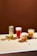 Four different beverages are arranged on a light-colored tabletop against a solid brown background. From left to right, there is a tall glass with a green drink and ice, a glass of red liquid with foam on top and a strawberry beside it, a creamy-looking beverage with orange chunks in a clear glass, and a mug with a yellow drink containing visible slices of fruit. Small fruits and nuts are scattered around the drinks.