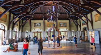 An expansive indoor space with high, intricate wooden beams and a large chandelier hanging from the ceiling. Large windows and doors allow natural light to flood the area. People are casually walking and interacting within the space, which also features display boards and art on the walls.