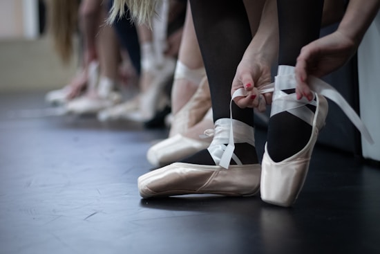 Several dancers are seated on the floor, preparing by tying the ribbons of their pointe shoes. The focus is on delicate hands adjusting the satin ballet shoes, with a sense of concentration and preparation for a performance or practice.