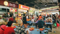 A bustling food court filled with people sitting at tables, eating and socializing. Several food stalls line the backdrop, displaying a variety of colorful signs with menus and food options. The seating area has blue tables and stools, while patrons in casual clothing enjoy their meals. Many people are wearing masks, creating a lively and vibrant atmosphere.
