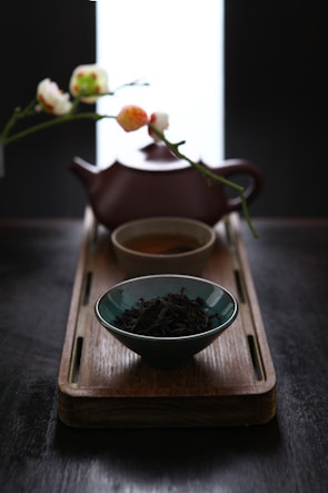 A wooden tray holds a blue bowl filled with loose black tea leaves, a small teacup with tea, and a brown teapot positioned in the background. Delicate flowers on a branch are visible behind the teapot, adding a touch of elegance to the scene.