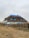 A traditional house with a thatched roof and blue accents sits behind a wooden fence. The building features multiple rounded windows and a blue chimney. The surrounding area is a dry, grassy field under an overcast sky.