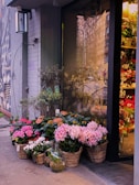 A charming flower shop entrance is adorned with a variety of colorful plants in woven baskets and pots. The assortment includes blooming pink and white flowers, adding a vibrant touch to the urban sidewalk setting. A glass vase with a small plant is placed among the baskets, enhancing the aesthetic. The shop's glass door reflects the street scene outside, including trees and buildings.
