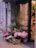 A charming flower shop entrance is adorned with a variety of colorful plants in woven baskets and pots. The assortment includes blooming pink and white flowers, adding a vibrant touch to the urban sidewalk setting. A glass vase with a small plant is placed among the baskets, enhancing the aesthetic. The shop's glass door reflects the street scene outside, including trees and buildings.