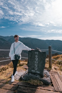 A person stands next to a large stone marker with Korean writing, set against a backdrop of mountains under a partly cloudy blue sky. The individual is casually dressed, leaning one arm on the marker. The ground is covered in dry grass and wooden planks, suggesting a hiking trail or lookout point.