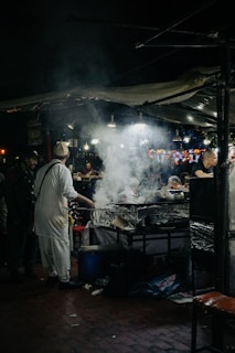 A bustling night market scene with a street food vendor preparing meals at a stall. Smoke rises from the cooking area, illuminated by several hanging lights. People can be seen interacting and waiting for food amidst the lively atmosphere.