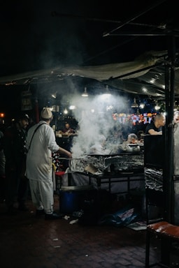 A bustling night market scene with a street food vendor preparing meals at a stall. Smoke rises from the cooking area, illuminated by several hanging lights. People can be seen interacting and waiting for food amidst the lively atmosphere.