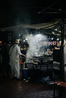 A bustling night market scene with a street food vendor preparing meals at a stall. Smoke rises from the cooking area, illuminated by several hanging lights. People can be seen interacting and waiting for food amidst the lively atmosphere.