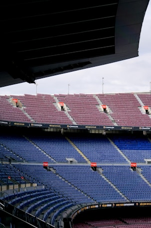 Rows of empty stadium seats in shades of blue and maroon fill the image. Overhead, part of the stadium roof is visible, casting shadows onto some of the seats. Along the edge of the seating tiers are banners and advertisements, with phrases such as 'Més Que Un Club' prominently displayed.