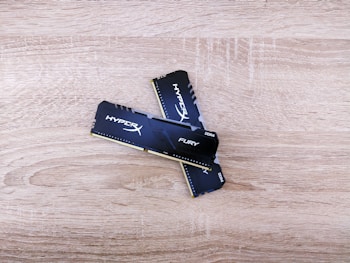 Two HyperX Fury DDR4 RAM sticks are placed on a wooden surface, slightly crossed over each other. The RAM sticks have a black exterior with white and silver branding.