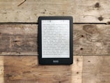 An e-reader displaying text in Russian is placed on a rustic wooden surface, which has varied tones and textures. The e-reader shows a page of text, and the overall setting gives a natural and relaxed vibe due to the weathered wood.