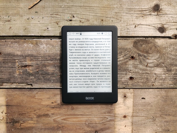 An e-reader displaying text in Russian is placed on a rustic wooden surface, which has varied tones and textures. The e-reader shows a page of text, and the overall setting gives a natural and relaxed vibe due to the weathered wood.