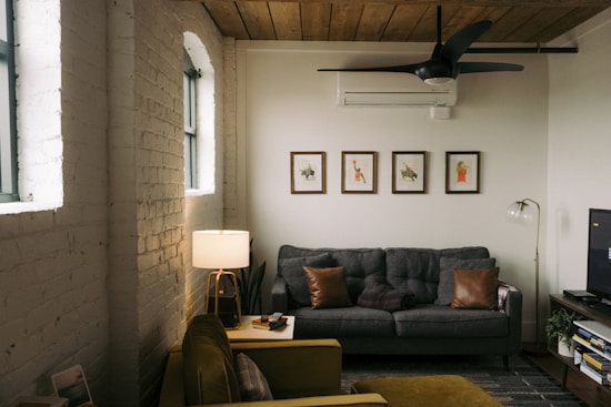 A cozy living room with a dark grey sofa adorned with brown cushions. Five framed art prints are arranged on the wall behind the sofa. A wooden ceiling with a modern black fan adds warmth to the space. To the left, a table lamp emits soft light, accenting the white brick wall with arched windows. An armchair is partially visible along with a side table holding magazines. The floor is covered with a subtle patterned rug.