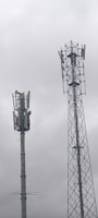 Two telecommunications towers are set against a cloudy sky. One tower has a solid cylindrical structure with various antennae attached, while the other tower has a lattice framework design with similar attachments.