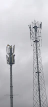 Two telecommunications towers are set against a cloudy sky. One tower has a solid cylindrical structure with various antennae attached, while the other tower has a lattice framework design with similar attachments.