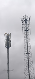 Two telecommunications towers are set against a cloudy sky. One tower has a solid cylindrical structure with various antennae attached, while the other tower has a lattice framework design with similar attachments.