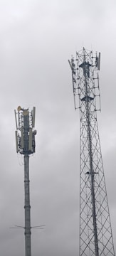 Two telecommunications towers are set against a cloudy sky. One tower has a solid cylindrical structure with various antennae attached, while the other tower has a lattice framework design with similar attachments.