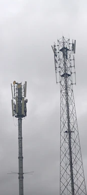 Two telecommunications towers are set against a cloudy sky. One tower has a solid cylindrical structure with various antennae attached, while the other tower has a lattice framework design with similar attachments.